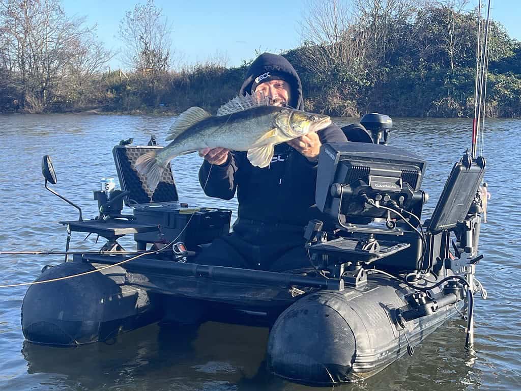 Een persoon in een kleine boot houdt een grote vis vast op een kalm wateroppervlak, omgeven door vegetatie.