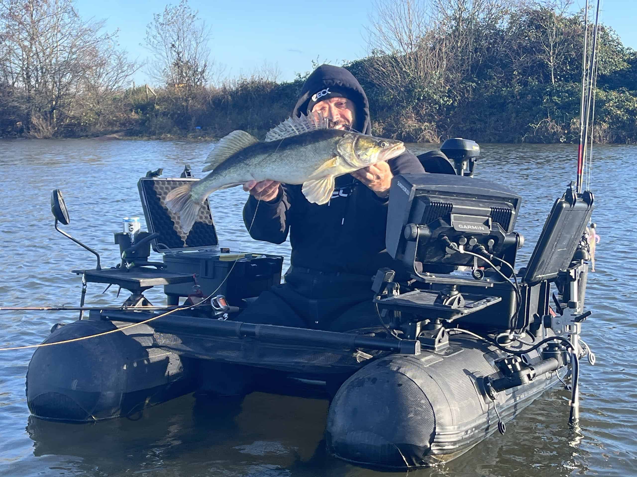 Een persoon die in een kleine opblaasbare boot op een rivier zit, houdt een grote vis vast. Visgerei en elektronica zijn zichtbaar op de boot. Bomen vormen de achtergrond.