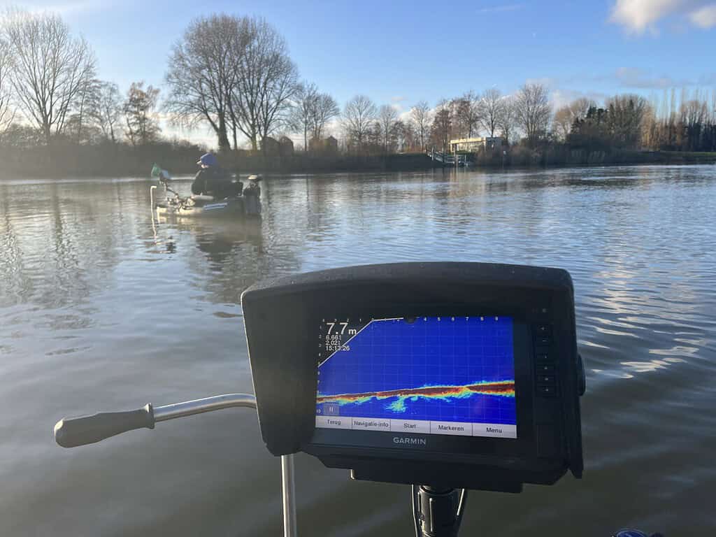 Boot met sonarweergave op een kalm meer, een andere boot in de verte, omringd door bladerloze bomen onder een blauwe lucht.