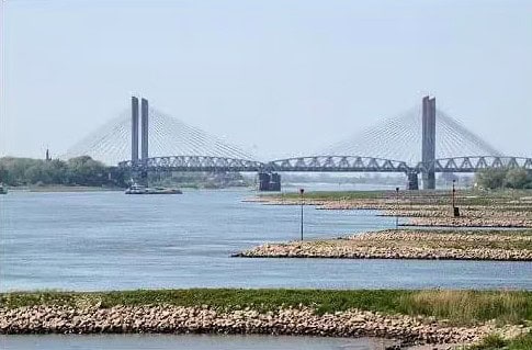 De tuibrug overspant sierlijk de Waal, waar de torenhoge structuur de rotsachtige oevers ontmoet. Onder de heldere hemel is hij veerkrachtig, zelfs tijdens hoogwater, een toevluchtsoord voor liefhebbers van feedervissen langs de serene wateren.