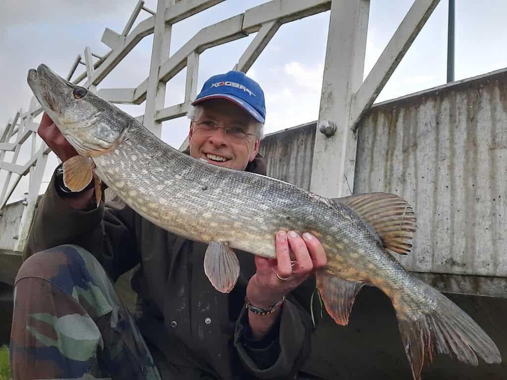 Krijn van Urk staat trots buiten bij een brug, met een grote vis in zijn handen die met vakmanschap is gevangen met Kunstaas. Het daglicht schittert op het water en benadrukt de prestatie van een perfecte dag vissen.