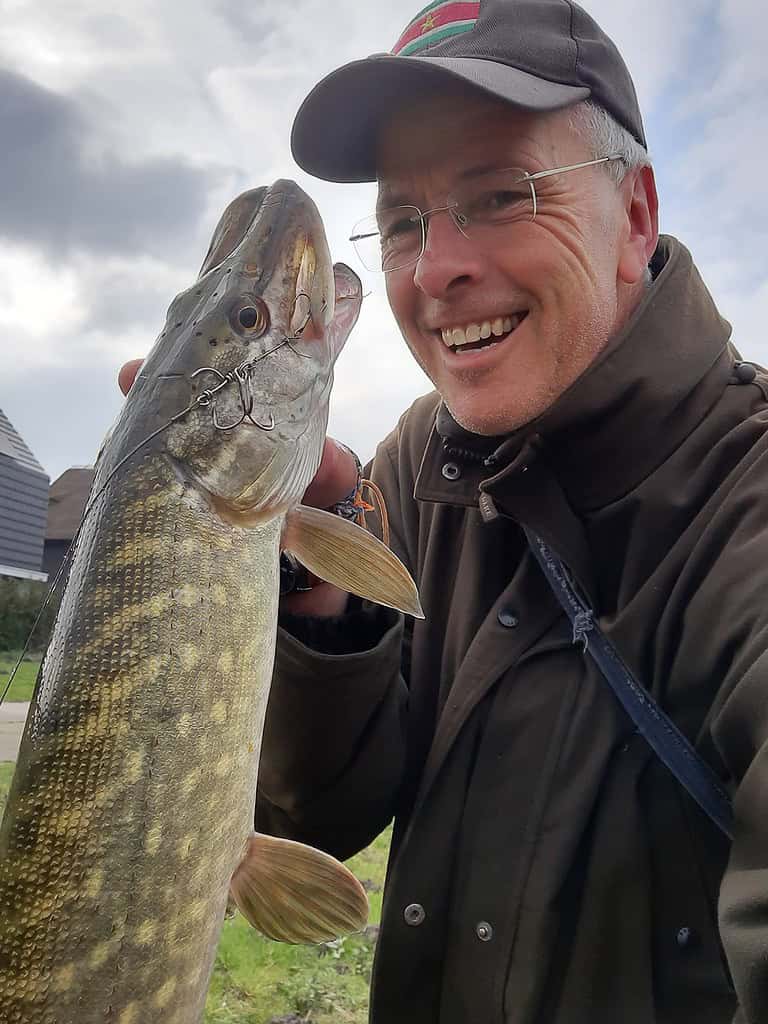 Krijn van Urk straalt breed, trots een grote vis buiten vasthoudend. Met een pet en bril op geniet hij duidelijk van zijn succesvolle Kunstaas-vangst.