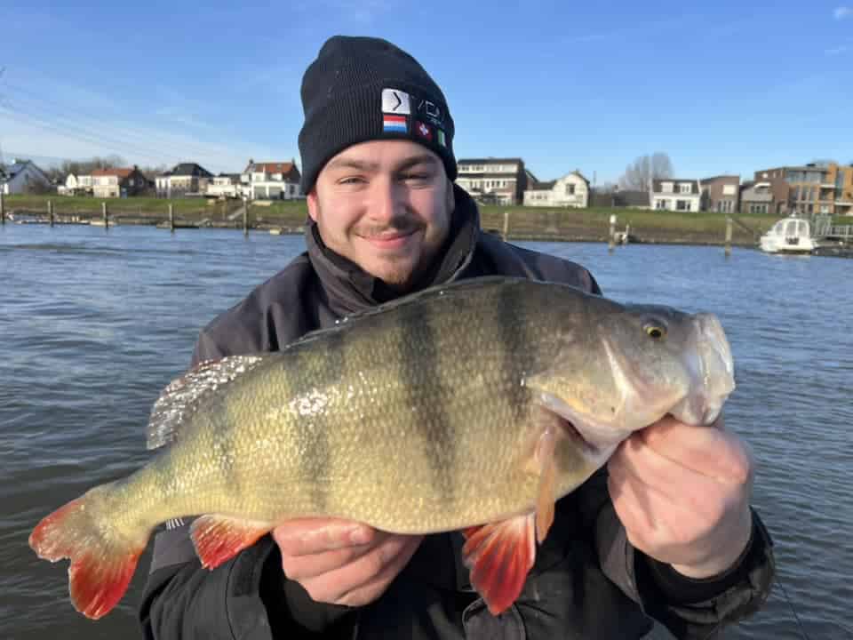 Een persoon houdt trots een grote vis vast, ter ere van het hoogtepunt van zijn visweek, met op de achtergrond een rivier en schilderachtige huisjes.