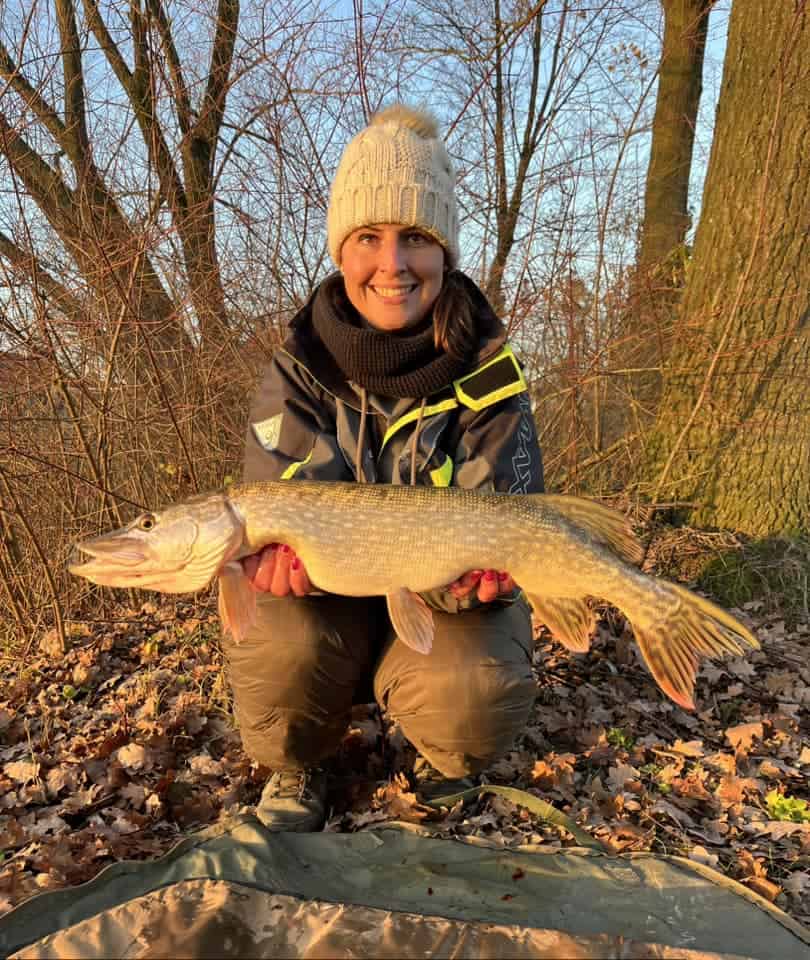 Een persoon in winterkleding toont trots zijn teleur van de week, een grote vis, tegen een achtergrond van bomen en gevallen bladeren.