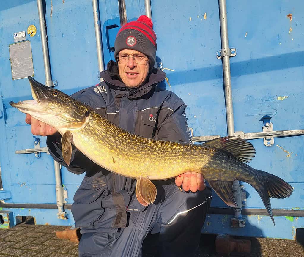 Persoon in een marineblauw jasje en een rode hoed toont trots zijn Vangsten van de Week, een grote vis, tegen de achtergrond van een blauwe zeecontainer.