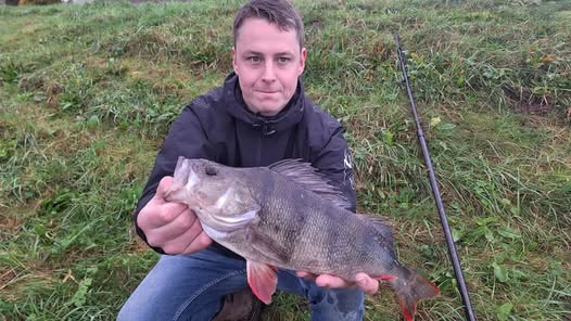 Persoon die trots een grote vis laat zien op de grasgrond, met een hengel in de buurt, wat doet denken aan de vangsten van de week.