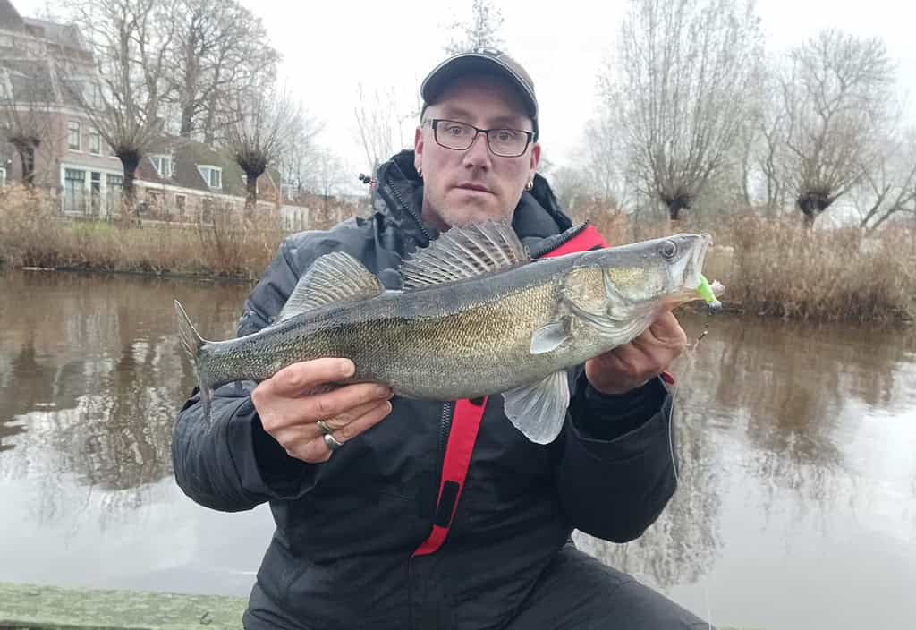 Een persoon toont trots zijn vangst aan de rivier, met een bril, een pet en een jas. Het was een van die dagen vol rustige vangsten, omringd door bomen en huizen op de achtergrond.