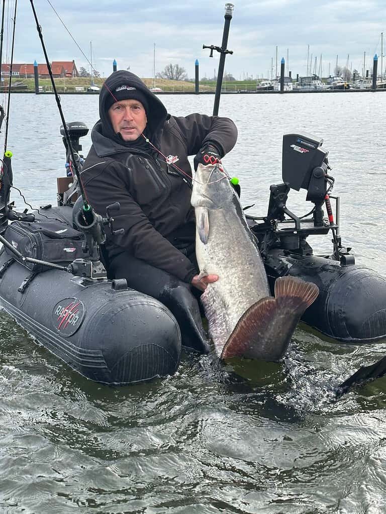 Een persoon zit in een opblaasboot en houdt met één hand trots een grote vangsten vast. Het water strekt zich achter hen uit en leidt naar de kustlijn bezaaid met gebouwen, wat het einde markeert van hun week vol avontuurlijke dagen vissen.