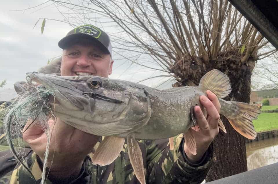 Een persoon in een camouflagejasje en met een pet lacht naar de camera en toont trots zijn vangsten, terwijl hij een grote vis vasthoudt tegen de achtergrond van weelderig groen.