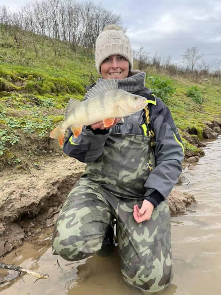 Een persoon in een camouflagewaadpak en een muts houdt trots een vis omhoog aan de oever van de rivier. Hij legt dit moment vast voor Vangsten van de week en lacht naar de camera.