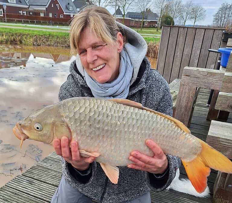 Zittend op een houten steiger aan de rivier, houdt een persoon trots een grote vis vast, hun nieuwste vangsten van de week. Op de achtergrond maken huizen en bladloze bomen het vredige tafereel compleet.