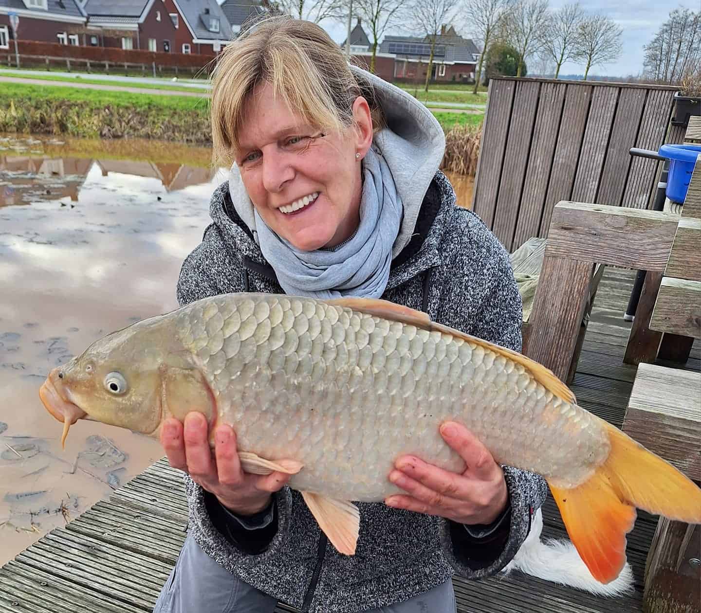 Zittend op een houten steiger aan de rivier, houdt een persoon trots een grote vis vast, hun nieuwste vangsten van de week. Op de achtergrond maken huizen en bladloze bomen het vredige tafereel compleet.