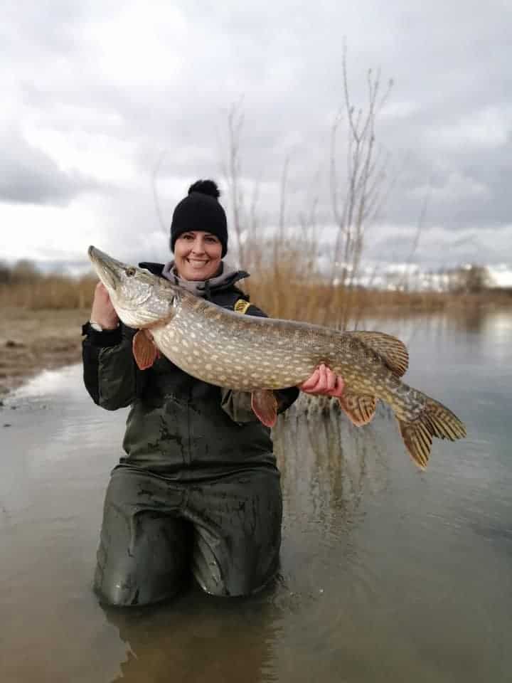 Een persoon knielt in het water, na een weeklange strijd als overwinnaar uit de strijd. Hij houdt een grote vangsten vast, met wuivend riet en een bewolkte lucht op de achtergrond.