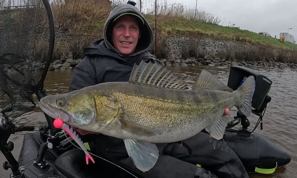 Een persoon in regenkleding, die zijn succesvolle vangsten van de week viert, houdt een grote vis met puntige vinnen vast. Ze zitten trots aan de oever van de rivier in een boot, omringd door visnetten en uitrusting, en reflecteren op hun triomfantelijke dag.