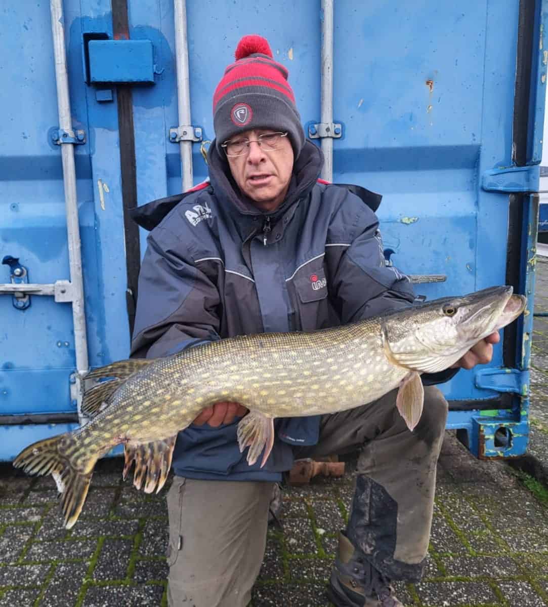 Een persoon met een muts en winterjas houdt triomfantelijk een grote vis buiten vast, staande voor een blauw metalen oppervlak. Deze vangst zal zeker worden gevierd als een van de vangsten van de week!.