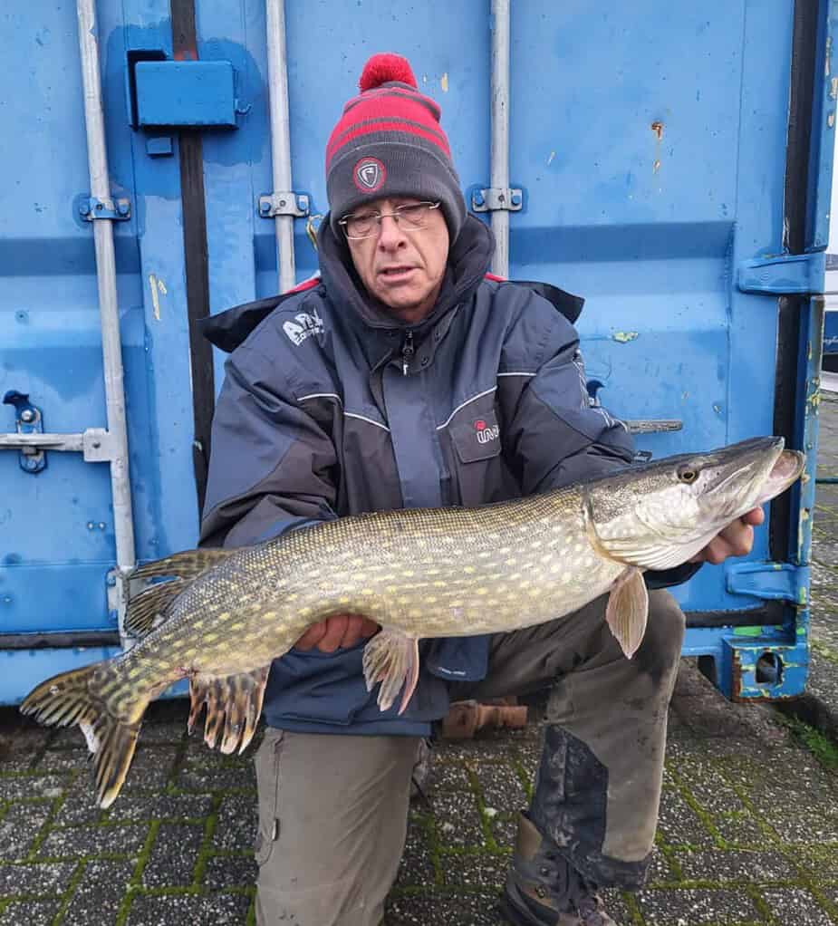 Een persoon toont trots zijn vangsten terwijl hij buiten een grote vis vasthoudt, gewikkeld in een winterjas en een muts.