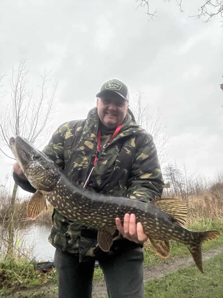 Een trotse visser, gekleed in een camouflagejack en pet, toont zijn vangsten (een grote vis) terwijl hij aan het glinsterende water staat.