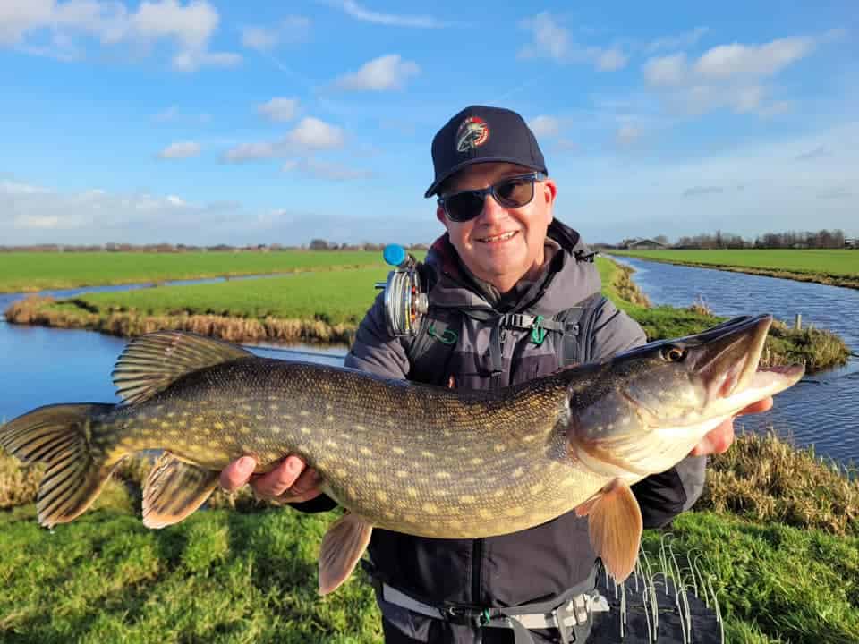 Een persoon in outdoorkleding toont trots zijn vangst tijdens de kersteditie. Hij houdt een grote vis vast aan de rivier op een zonnige dag, met een weelderig graslandschap op de achtergrond.