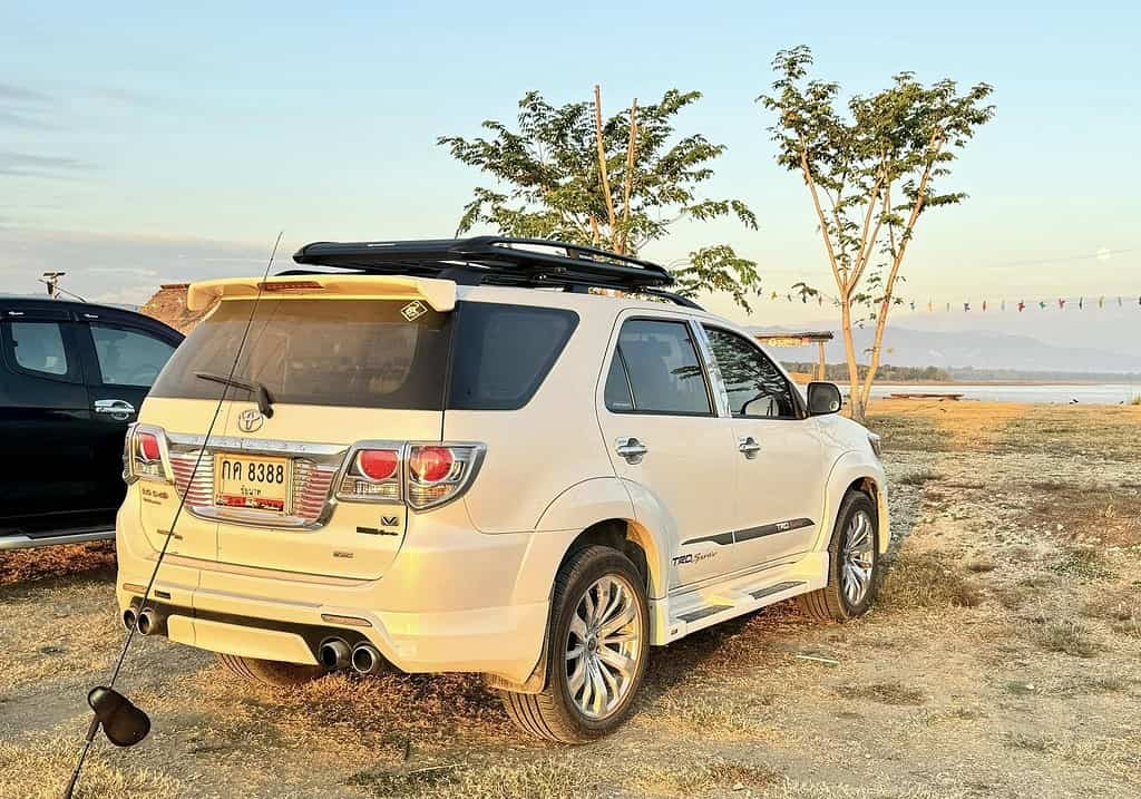 Een witte SUV staat geparkeerd op een grasveld bij een waterpartij, met bomen en in de verte een tent onder de heldere hemel van Thailand. De lokale bevolking gaat hier vaak vissen.