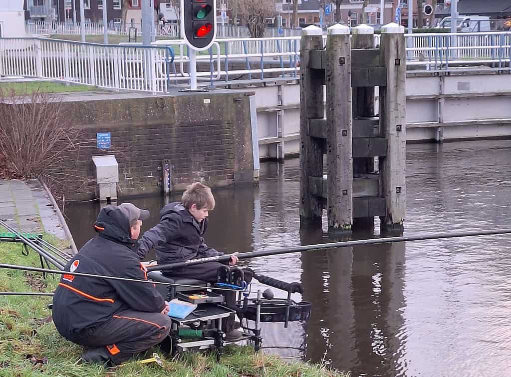 Twee mensen vissen aan een kanaal, zittend op het gras met hun spullen. Op de achtergrond zijn een brug en een stoplicht zichtbaar, wat doet denken aan een serene Talentenclinic-sessie geïnspireerd door Stefan Hooymans witvissen-expertise.