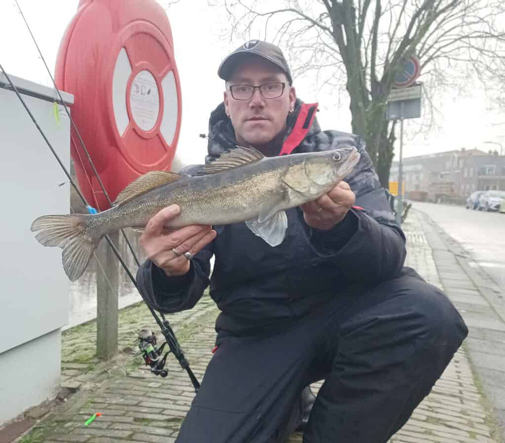 Knielend op de stoep met een vangst, toont een persoon trots zijn vis naast een rode reddingsboei en hengel. Het is een indrukwekkende vangsten, terwijl bomen en gebouwen de perfecte achtergrond vormen.