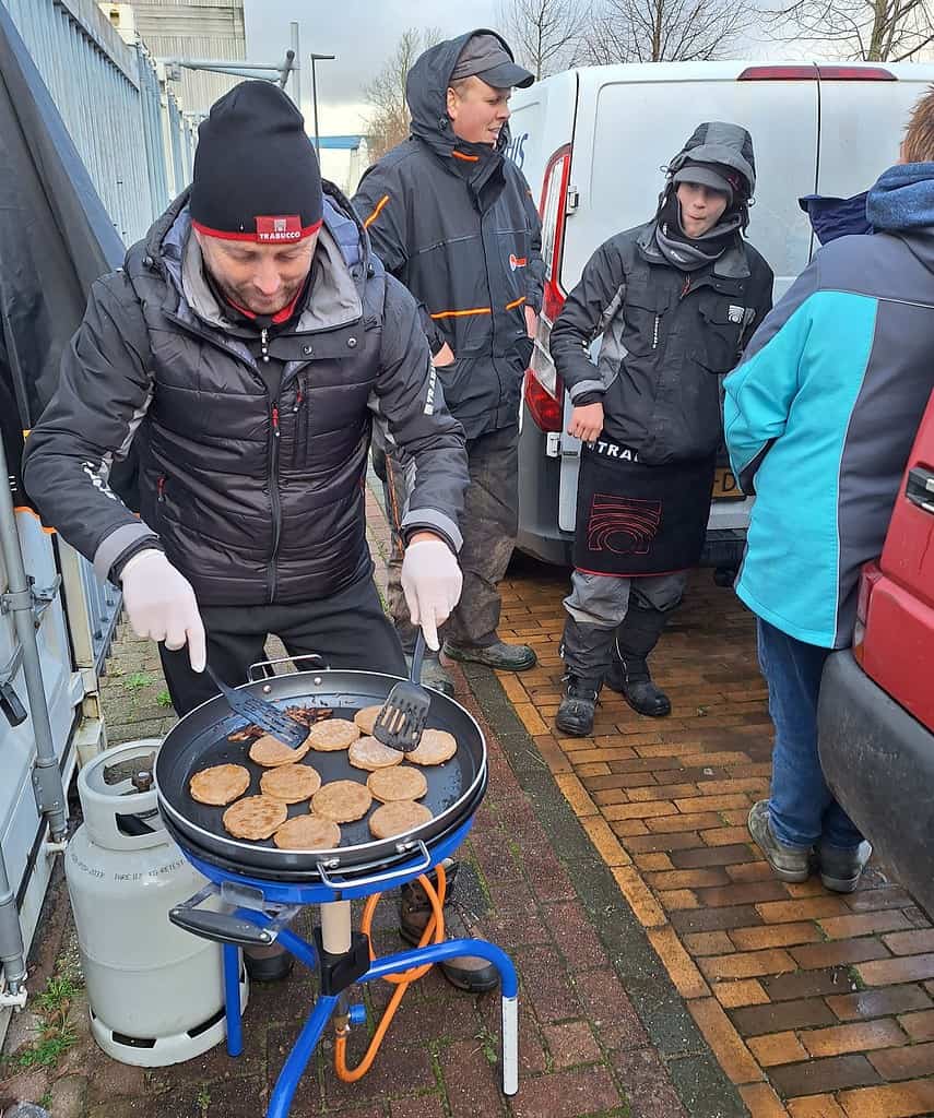 Terwijl Stefan Hooyman vakkundig hamburgers grilt op een draagbare grill buiten, staan er drie anderen in de buurt, ingepakt in jassen en mutsen tegen het koude weer.