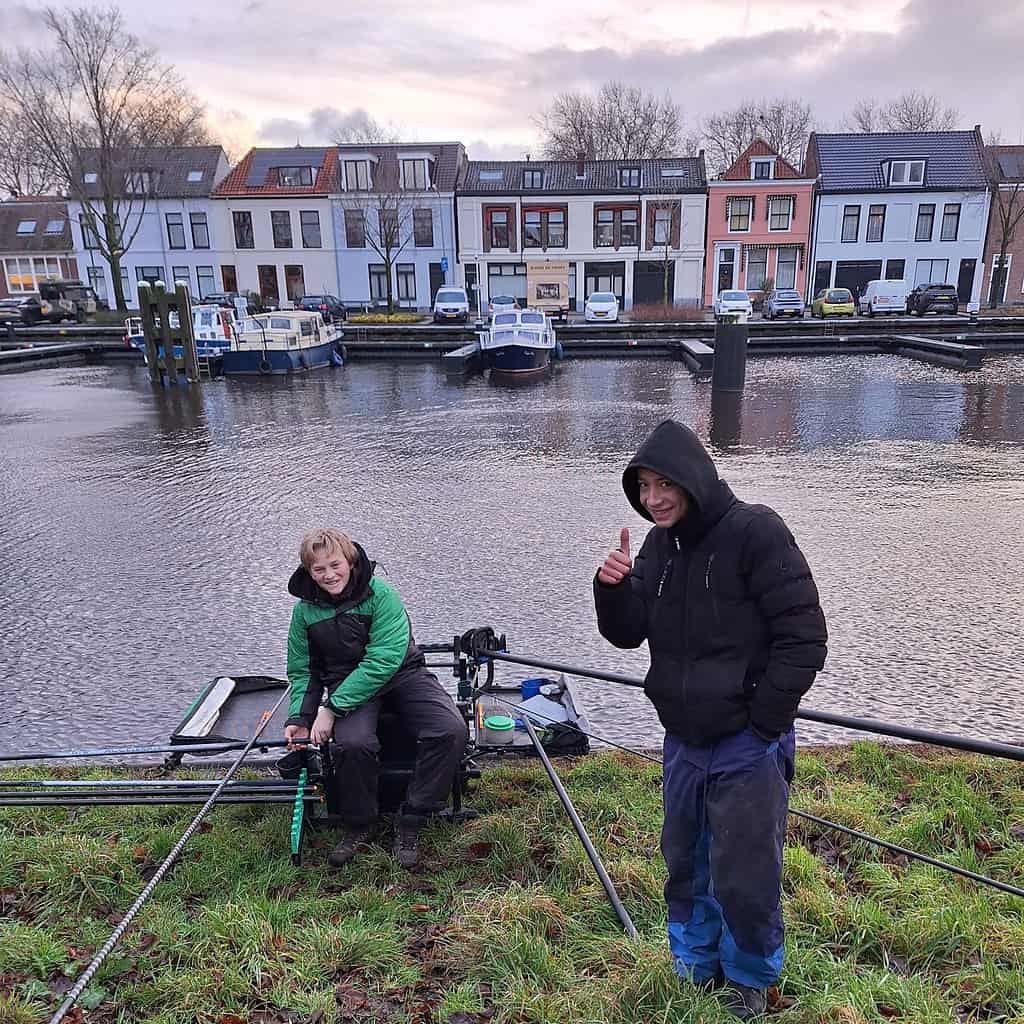 Aan een kanaal, waar kleurrijke kanaalboten en charmante huizen het decor vormen, houden twee vrienden zich bezig met witvissen. De een zit tevreden met visgerei en een warme glimlach, terwijl de ander, misschien geïnspireerd door de technieken van Stefan Hooyman, trots een duimpje omhoog steekt.