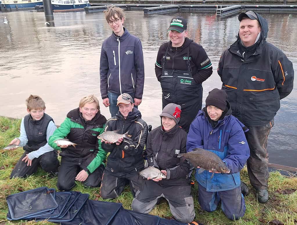 Een groep mensen, waaronder Stefan Hooyman, poseert bij het water met verschillende vissen die ze hebben gevangen tijdens een Talentenclinic. Ze hebben een brede glimlach op hun gezicht, zijn gekleed in outdoorkleding en laten hun witvissen skills zien.