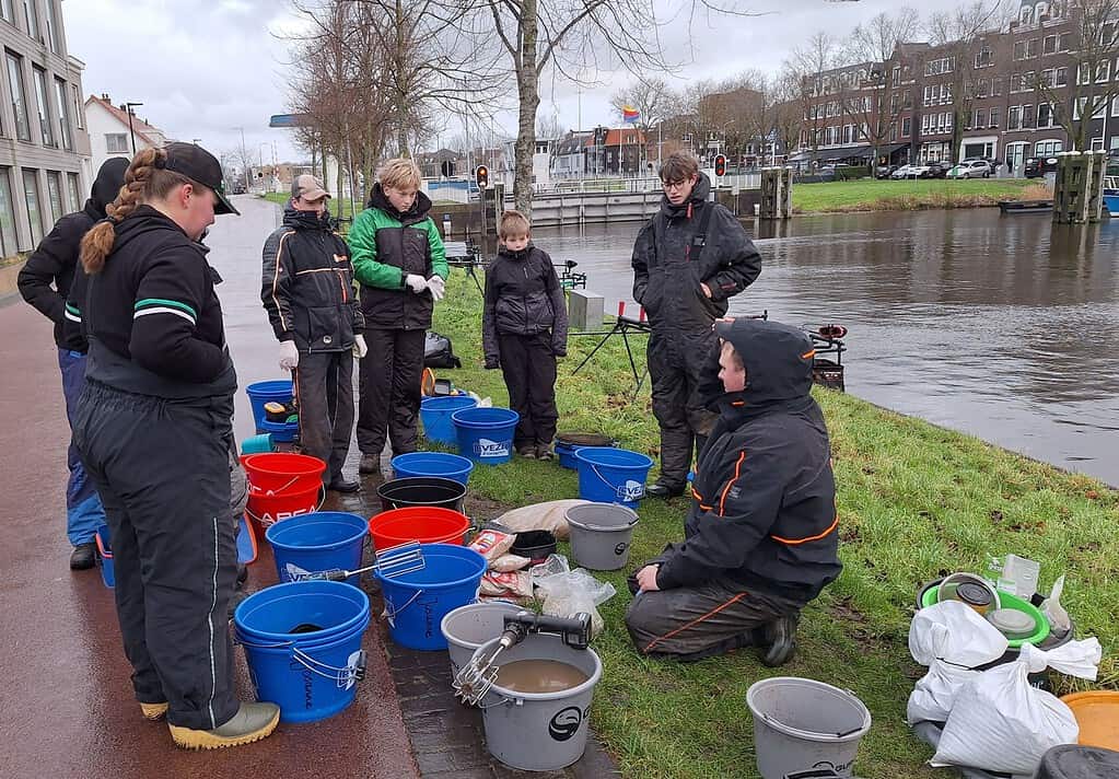 Een groep mensen in outdoorkleding verzamelt zich rond emmers en uitrusting aan de oever van een rivier in een stedelijk gebied. Stefan Hooyman knielt en lijkt witvissentechnieken uit te leggen of te demonstreren als onderdeel van de Talentenclinic aan de anderen.