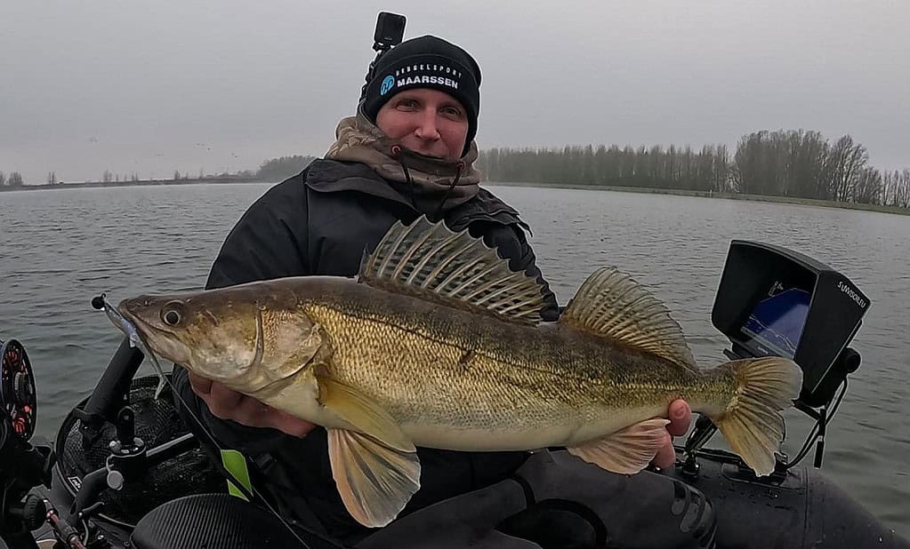 Een persoon in een boot houdt trots zijn vangst van de week vast, een grote vis, terwijl hij winterkleding draagt en een camera op zijn hoofd draagt. De achtergrond toont een sereen meer onder een bewolkte lucht, perfect om het hele jaar door vangsten vast te leggen.