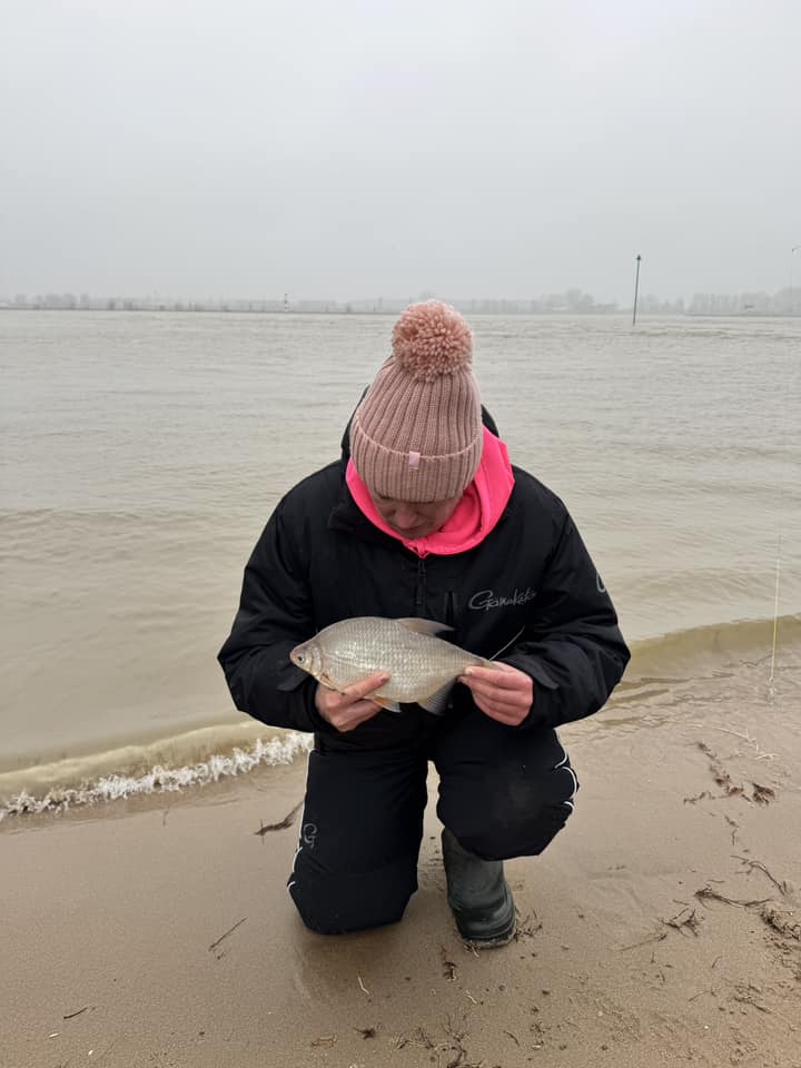 Een persoon knielt op het zandige strand en houdt trots een vis vast in zijn vangsten. De bewolkte lucht en het water vormen het decor van een onvergetelijk jaar.