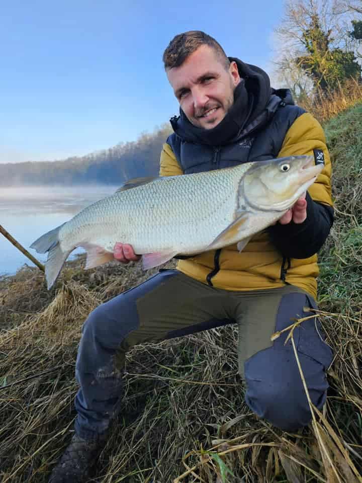 Een persoon knielt op de oever van de rivier en toont trots de vangst van de week. Hij houdt daarbij een grote vis met beide handen vast.