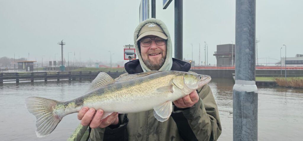 Een persoon met een hoodie en een bril toont trots zijn vangsten, een grote vis, terwijl hij bij een watermassa staat, met op de achtergrond industriële gebouwen.