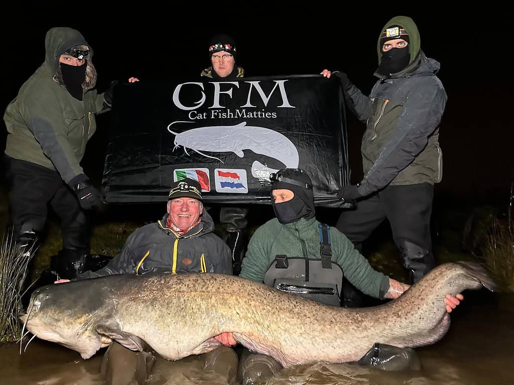 Vier mensen met een spandoek met de tekst "Cat Fish Matties" staan achter een persoon die knielt in het water met een grote meerval.