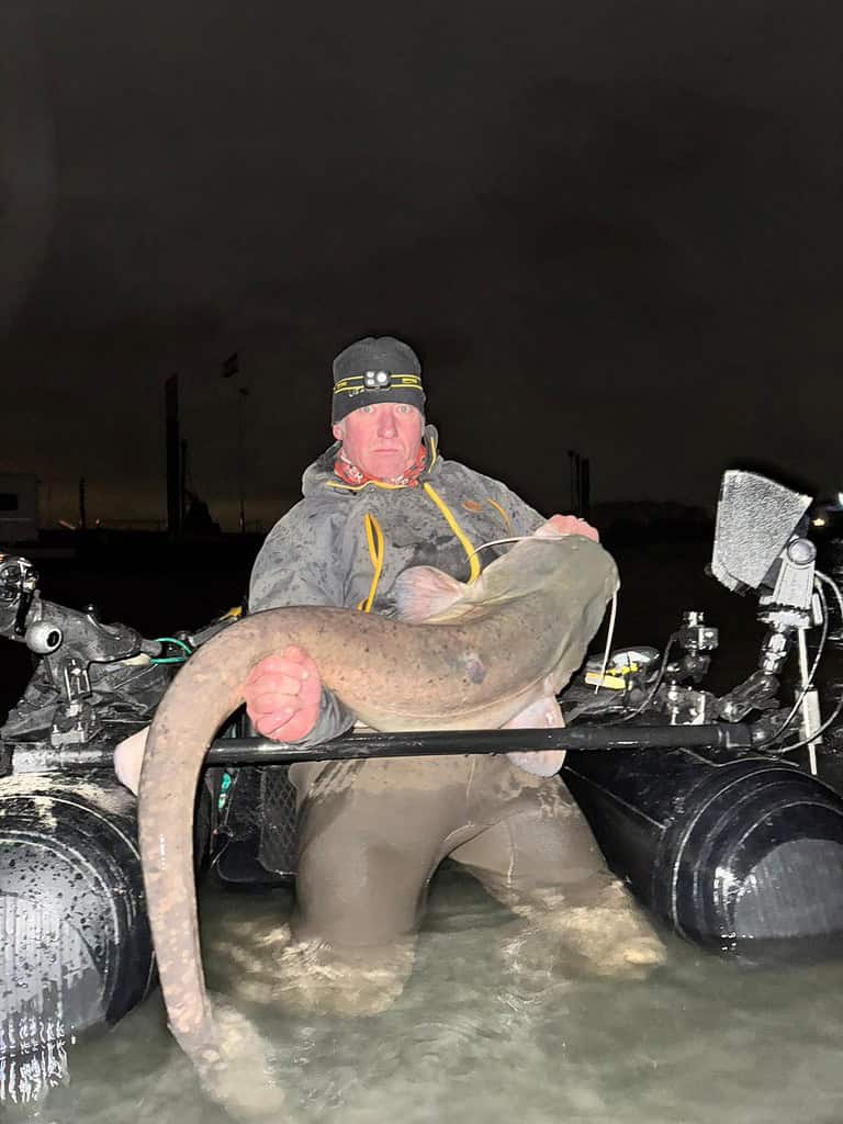 Een persoon die 's nachts in een kleine opblaasbare boot in het water staat, tot aan zijn middel, en een grote vis vasthoudt.