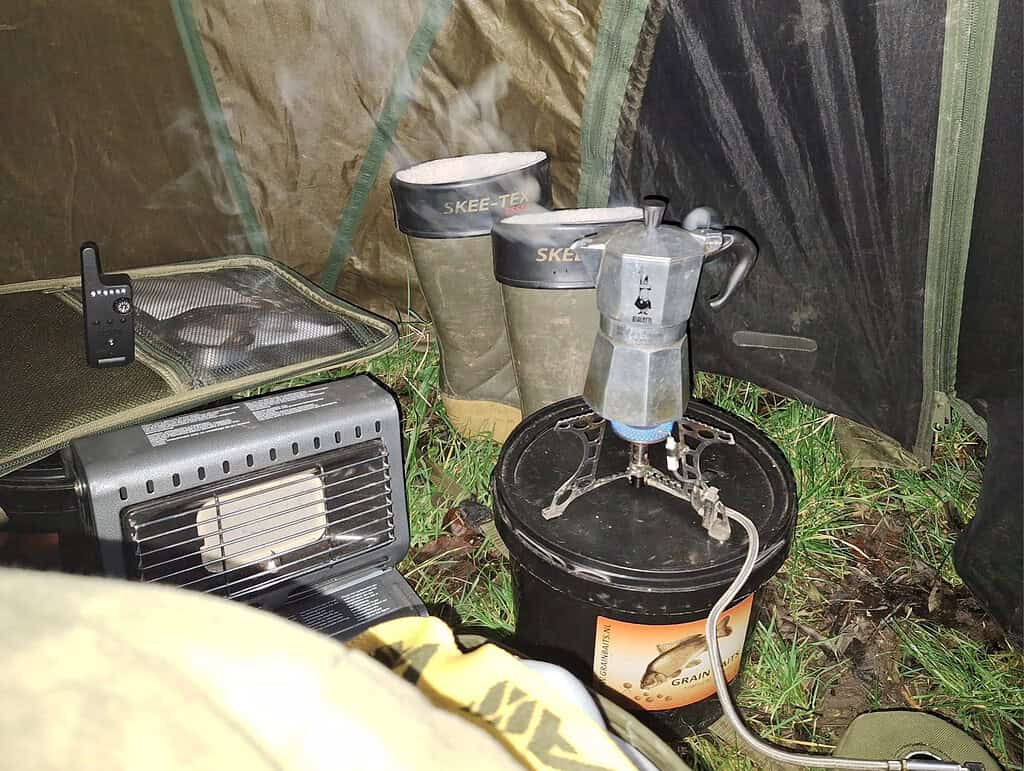 Kampeeruitrusting met een klein verwarmingselement, laarzen en een koffiepot op een gasbrander op een emmer in de tent, ideaal om op te warmen na een koude winterdag met karpervissen.