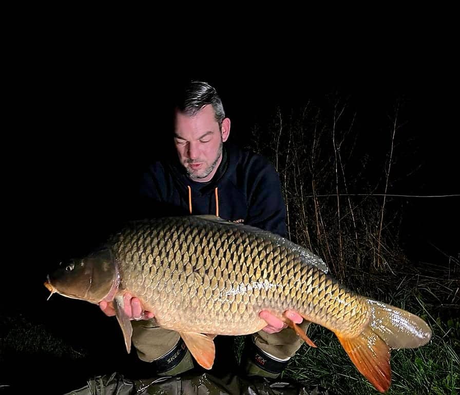 Een persoon die trots een grote karper vasthoudt onder de maanverlichte winterhemel, staand op het bevroren gras.