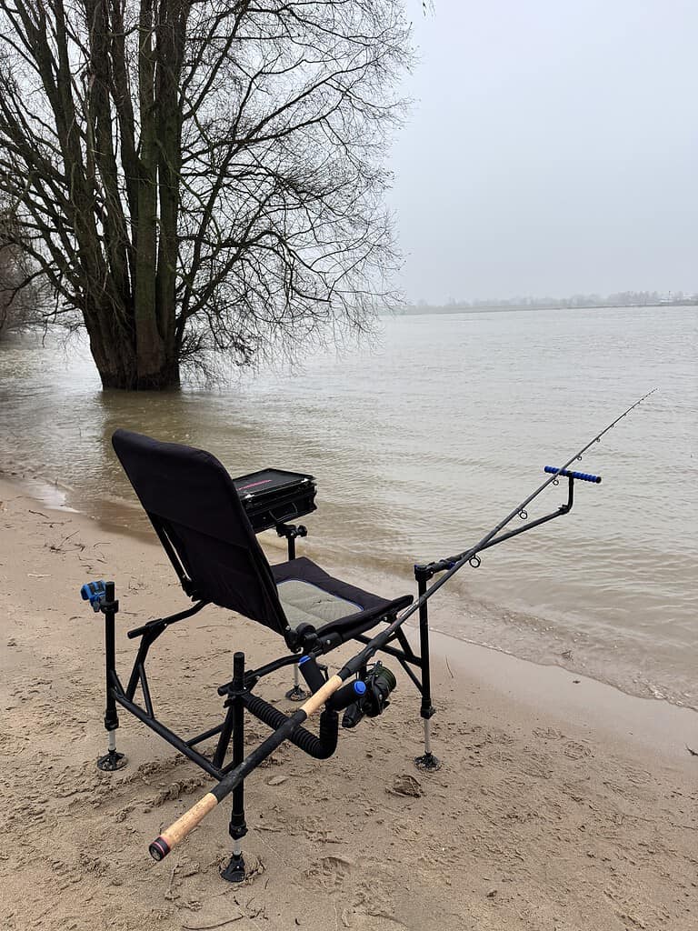 Op de zanderige oever van de rivier, naast een boom op een bewolkte dag, staat een visstoel met een hengel en een viskoffer. Dit is de perfecte plek om te witvissen.