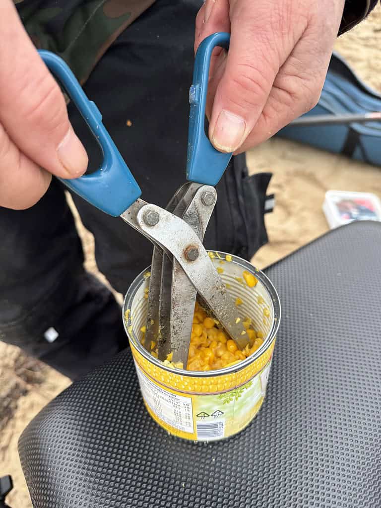 Handen die met een schaar met blauwe handvatten een blik maïs openen aan de Grote Rivier. Het gestructureerde oppervlak geeft dit buitentafereel een rustieke charme.