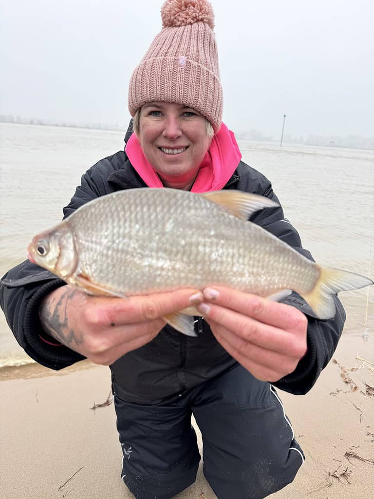 Knielend op een zandstrand, toont een persoon gekleed in een roze hoed en capuchonjack trots zijn vangst van witvissen langs de grote rivier. Op de achtergrond glinstert het water onder een bewolkte lucht, wat een sereen hoogwatertafereel creëert.