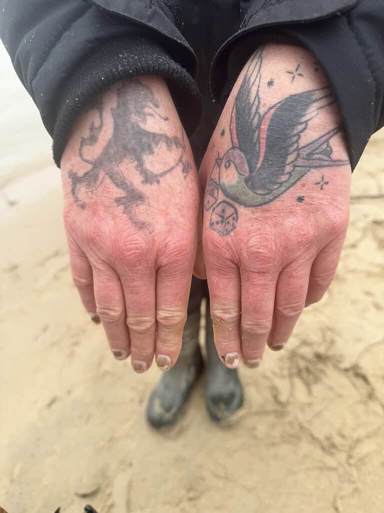 Een paar handen toont ingewikkelde tatoeages: een leeuw links en een vogel met dobbelstenen rechts. Het individu staat bij de grote rivier tijdens hoogwater, draagt een donker jasje en laarzen, hun voeten stevig geplant in het zandoppervlak.