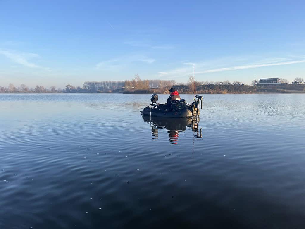 Een persoon drijft in een klein bootje op een kalm meer onder een helderblauwe lucht, met bomen aan de horizon net buiten bereik. Het lijkt alsof een kunstenaar de rust schildert op twee meter afstand van de natuur.