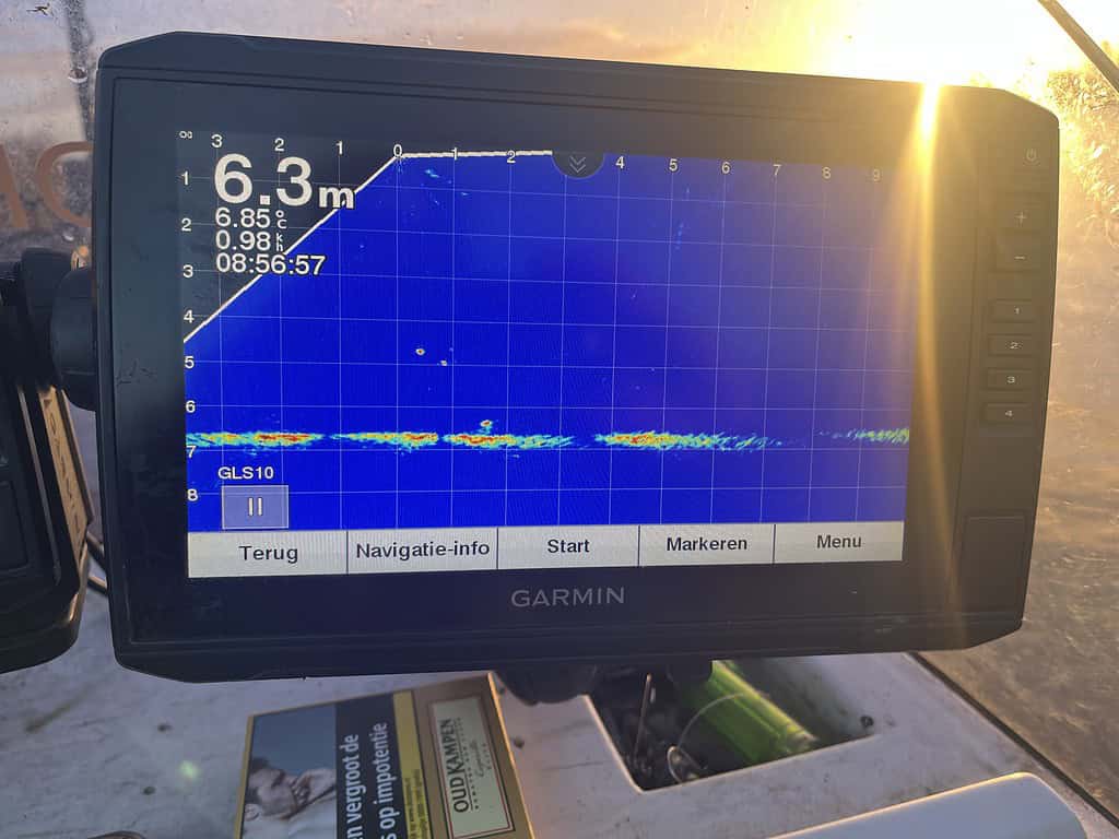 Een Garmin marine display toont dieptemetingen en sonargegevens op een raster, terwijl zonlicht gedeeltelijk op het scherm schijnt. Na een luie dag op zee biedt het een moment van helderheid vergelijkbaar met een goed gesprek met Roelof Hemmen.