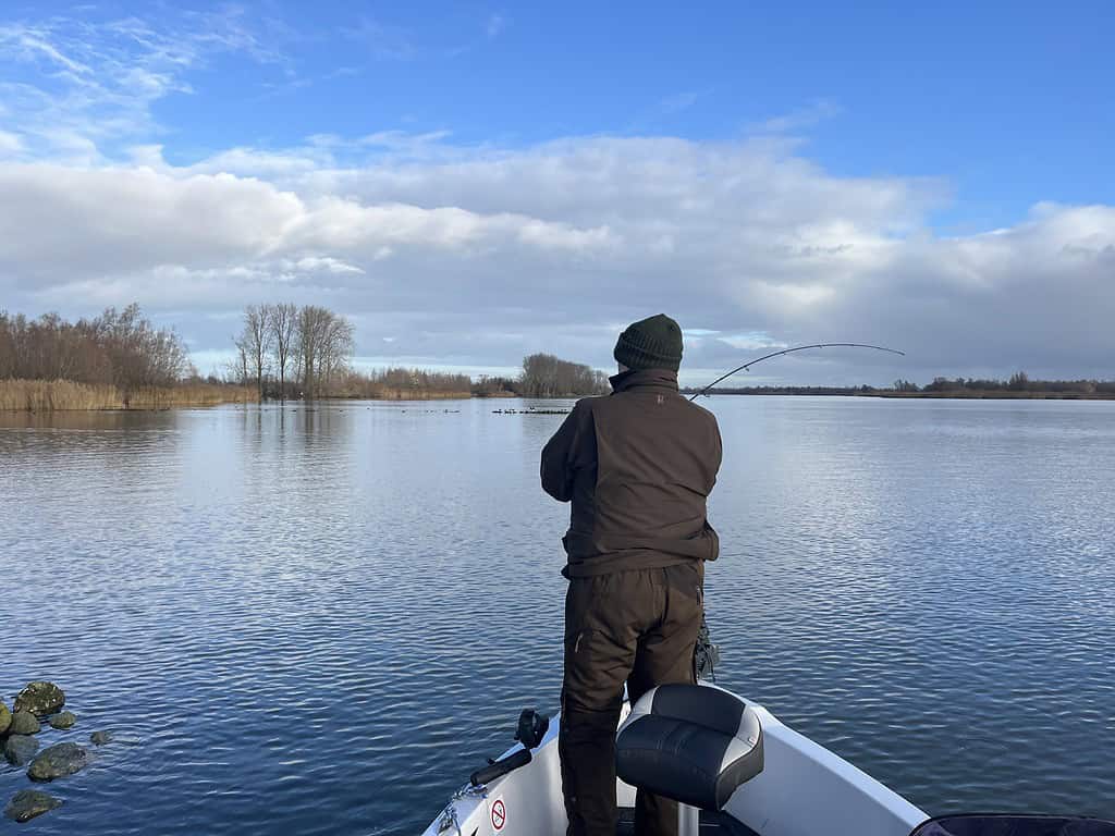 Persoon in een bruin jasje staat aan de rand van een boot op een loeitaaie dag, vissend op een kalm meer onder een gedeeltelijk bewolkte lucht. Bomen omlijnen de horizon terwijl Roelof Hemmen's stem echoot met goede gesprekken.