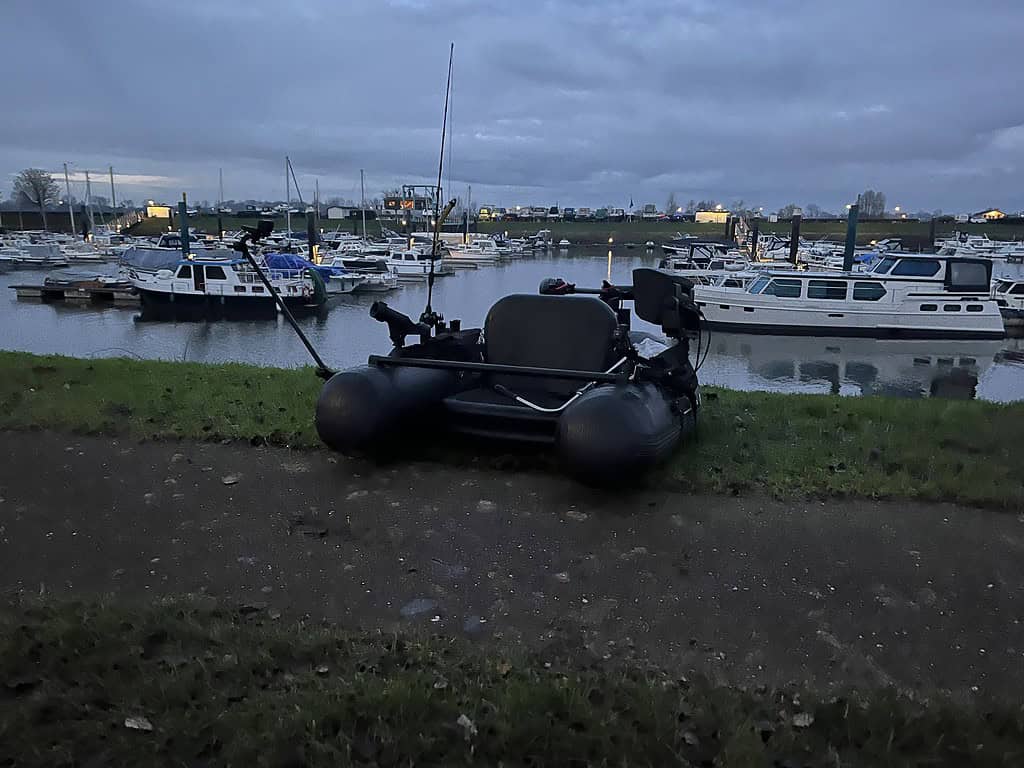 Opblaasbare boot met hengels en uitrusting op het gras naast een haven. In de schemering zijn verschillende aangemeerde boten zichtbaar.