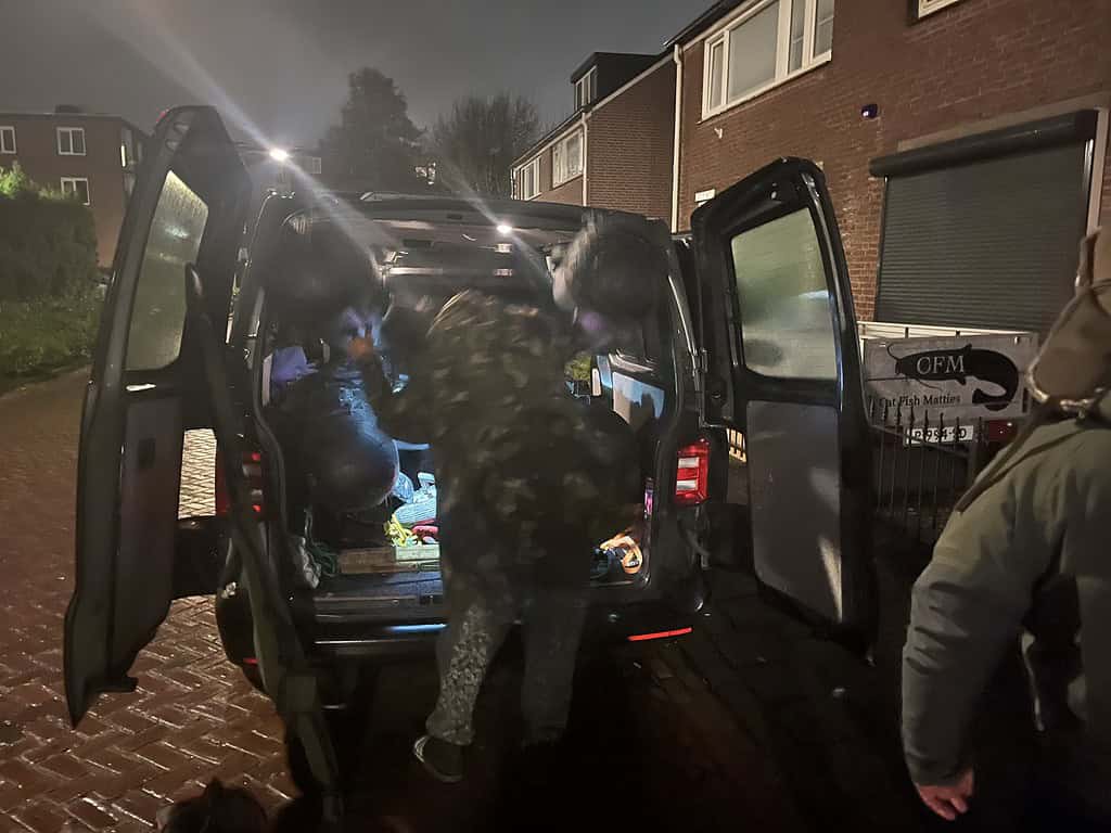 Drie mensen in camouflagejassen laden 's nachts spullen uit de laadbak van een busje dat geparkeerd staat op een natte weg.