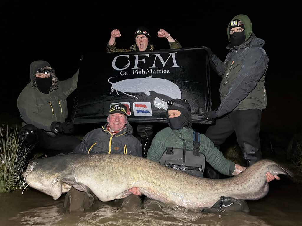 Vijf mensen in winterkleding houden een grote vis vast terwijl ze 's avonds in het water poseren, met op de achtergrond een spandoek met de tekst "CFM Cat Fish Matties", dat trots de vangsten van de week laat zien.