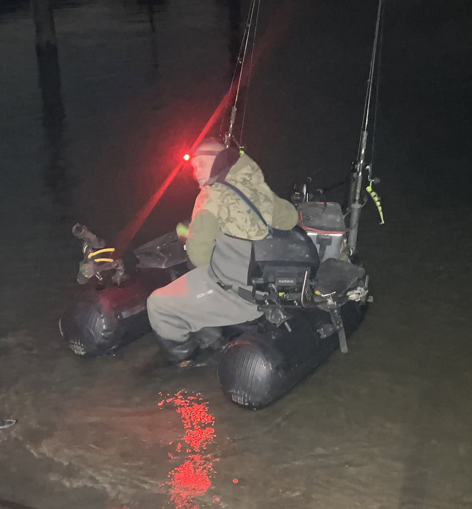 Persoon zit op een klein opblaasbaar vaartuig in donker water, draagt een hoofdlamp en winterkleding, met visuitrusting aan boord.