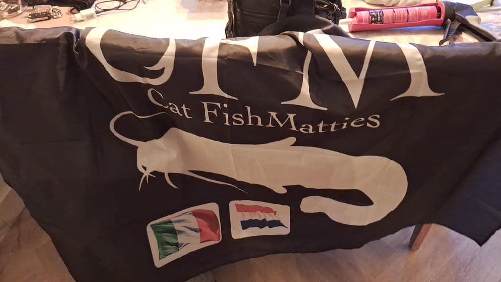 Een donkergekleurde banner met de tekst "GFM Cat Fish Matties", een afbeelding van een meerval en drie vlaggen: Italië, Nederland en een gedeeltelijk zichtbare vlag.