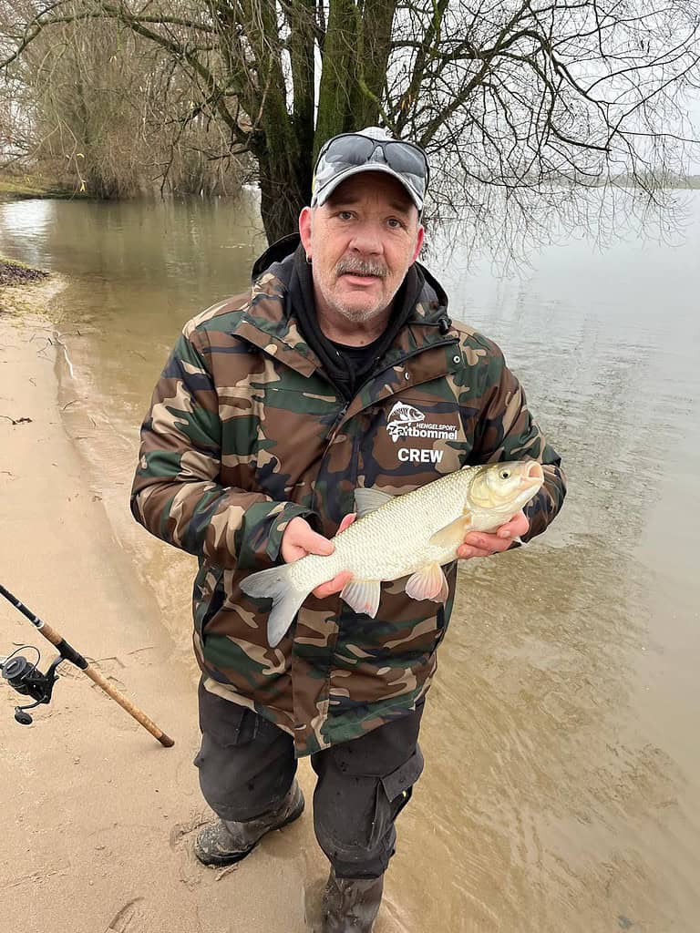 Een man in een camouflagejack houdt trots een vis vast aan de oever van een grote rivier, met zijn hengel op de grond, en legt de sensatie vast van witvissen tijdens hoogwater.